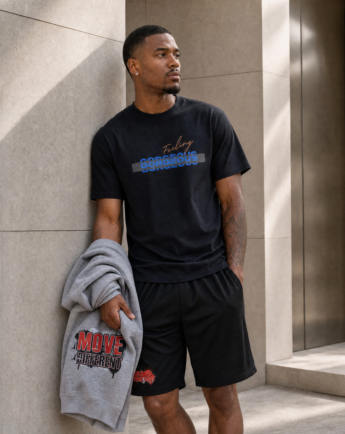 A man stands against a beige stone wall wearing a black Gorgeous Strength t-shirt and black Built to Stand Out shorts, holding a gray Signature Hoodie with red and black graphics.