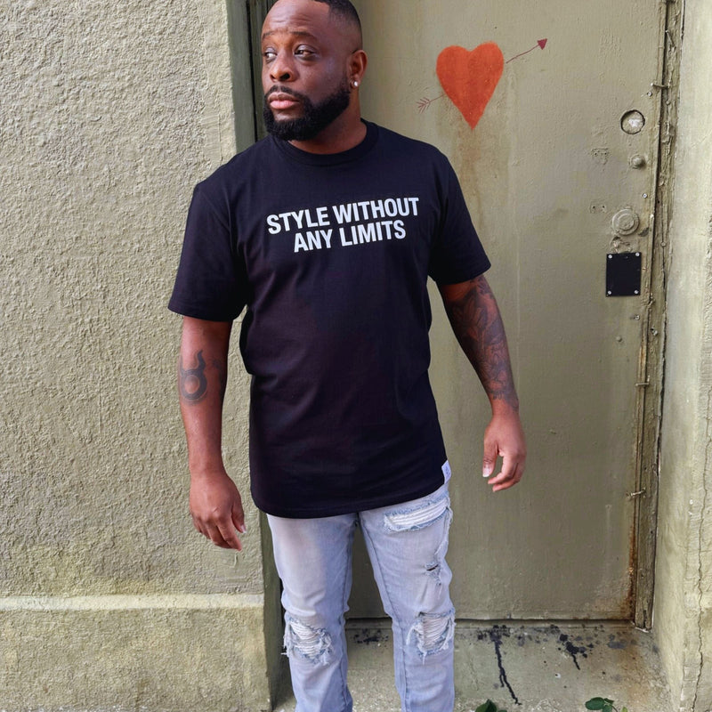 Man wearing a black t-shirt with 'STYLE WITHOUT ANY LIMITS' text, standing against a textured wall.