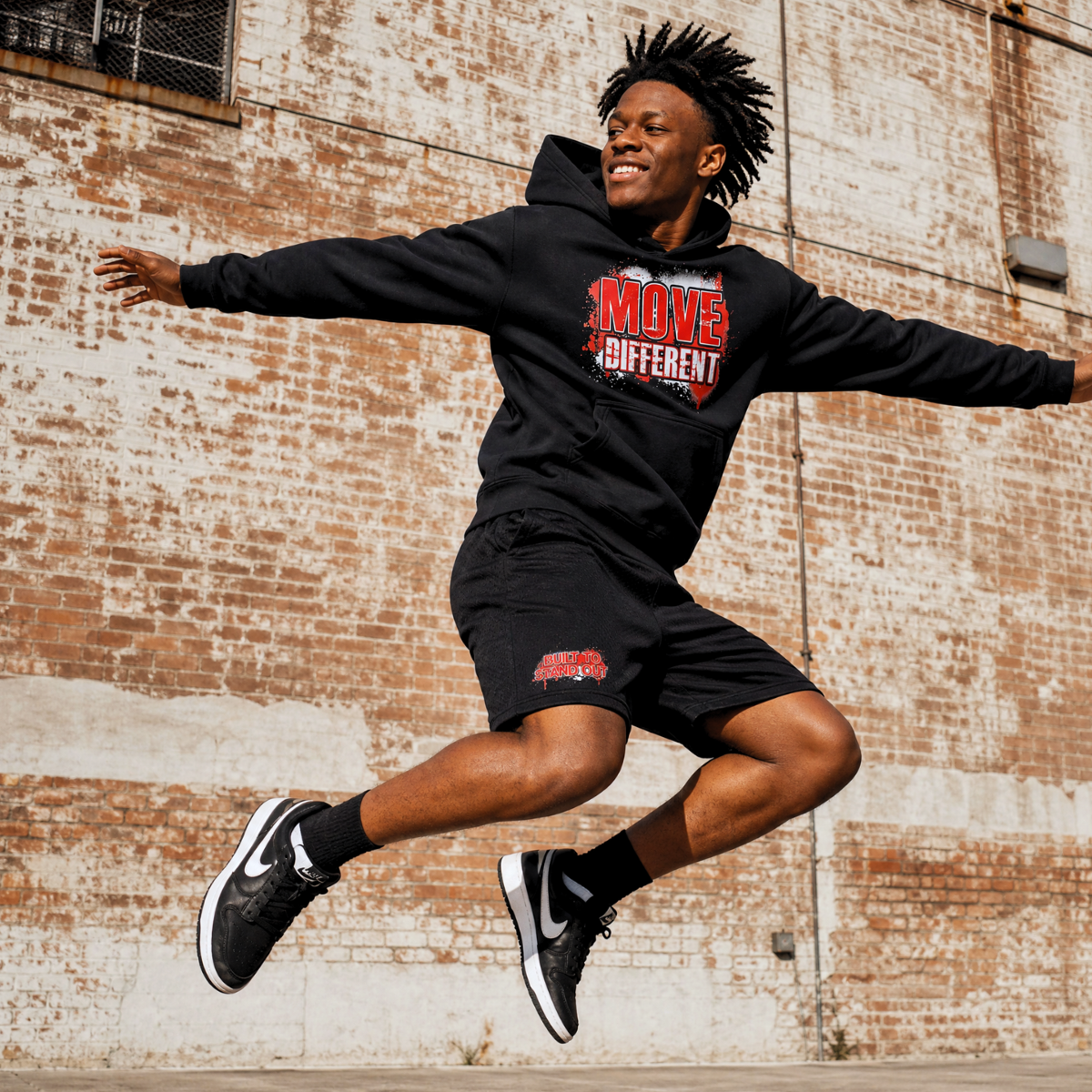 Person wearing a black My Gorgeous Clothing hoodie and shorts while jumping in an urban setting.